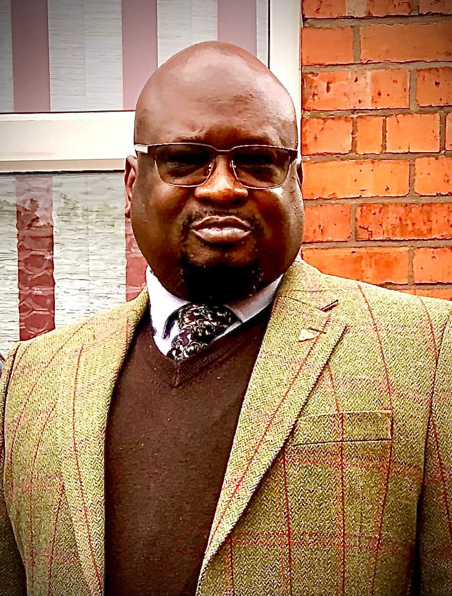 Bald Black man wearing glasses, a tweed blazer, and tie before a brick wall.